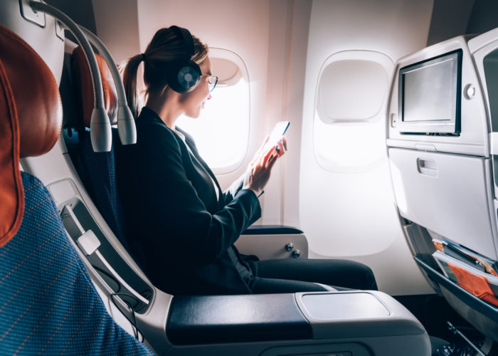 Woman sitting in window seat on airplane listening to media on her phone using noise cancelling headphones