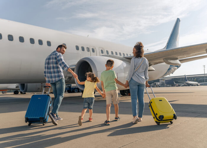 Parents and children walking toward plane on tarmac while wheeling suitcases