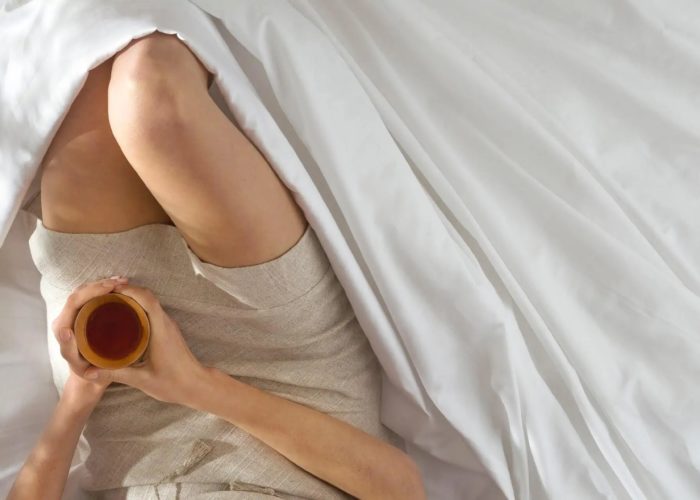 Overhead view of the legs of a person sitting in a pile of sheets and holding a cup of coffee